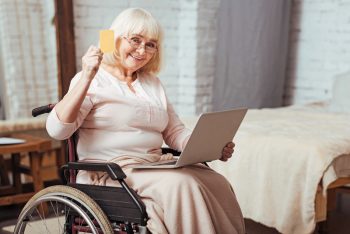 Older woman in wheelchair with laptop, cheerfully waving her credit card.