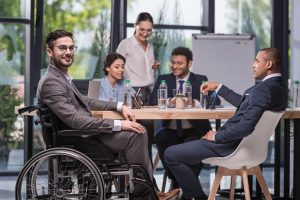 Diversity and inclusion example photo: A group of people in an office working at a table. In the foreground, separate from teh group, is a man in a wheelchair smiling at the camera. He does not appear to be part of the group. 