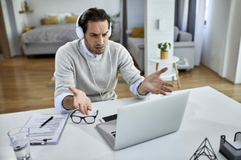 frustrated blind man in headphones gesturing irritably at his laptop.